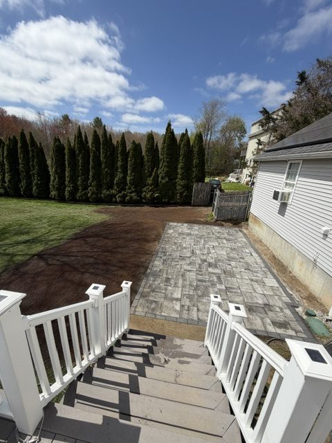 Gray paver patio beside a home with a row of arborvitae along the property line